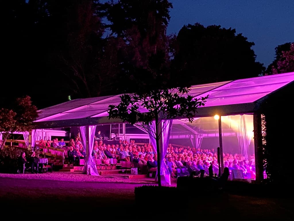 Temporary theatre marquee at night with illuminated stage and seated audience.